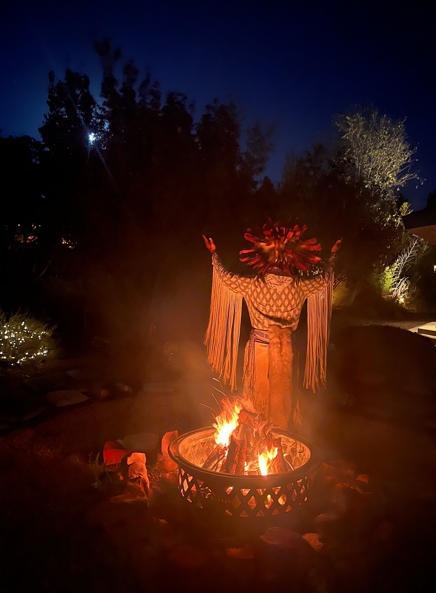 A ceremonial figure standing with raised arms behind a glowing fire at night