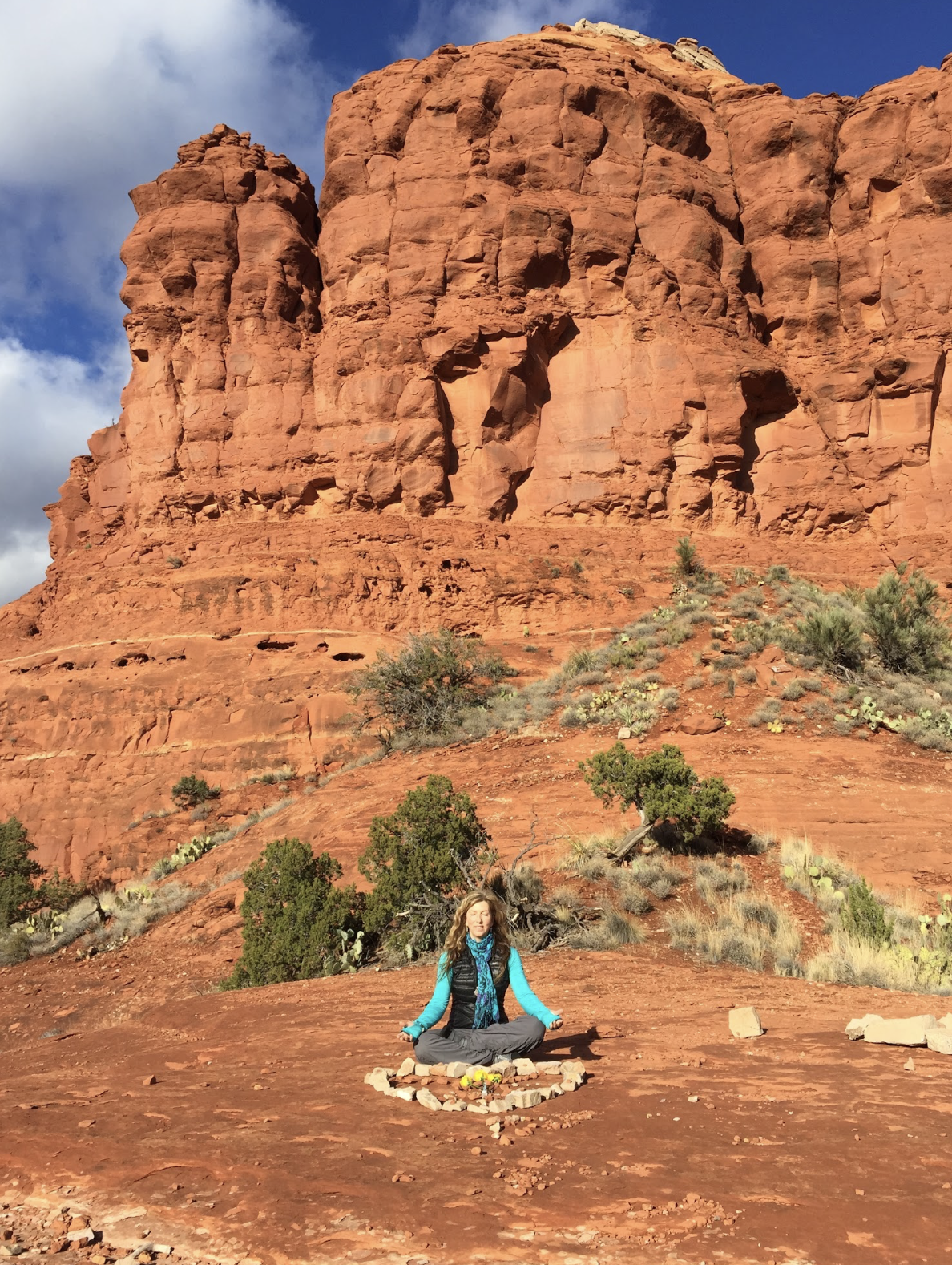 A woman meditating on red earth beneath a monumental sunlit Sedona cliff